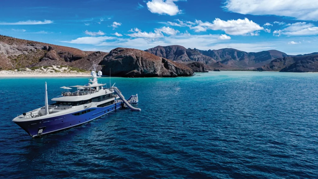 The luxury expedition yacht NOMAD anchored near the turquoise waters and white sand beaches of Balandra Bay in Baja California Sur, surrounded by dramatic rocky cliffs and a clear blue sky.